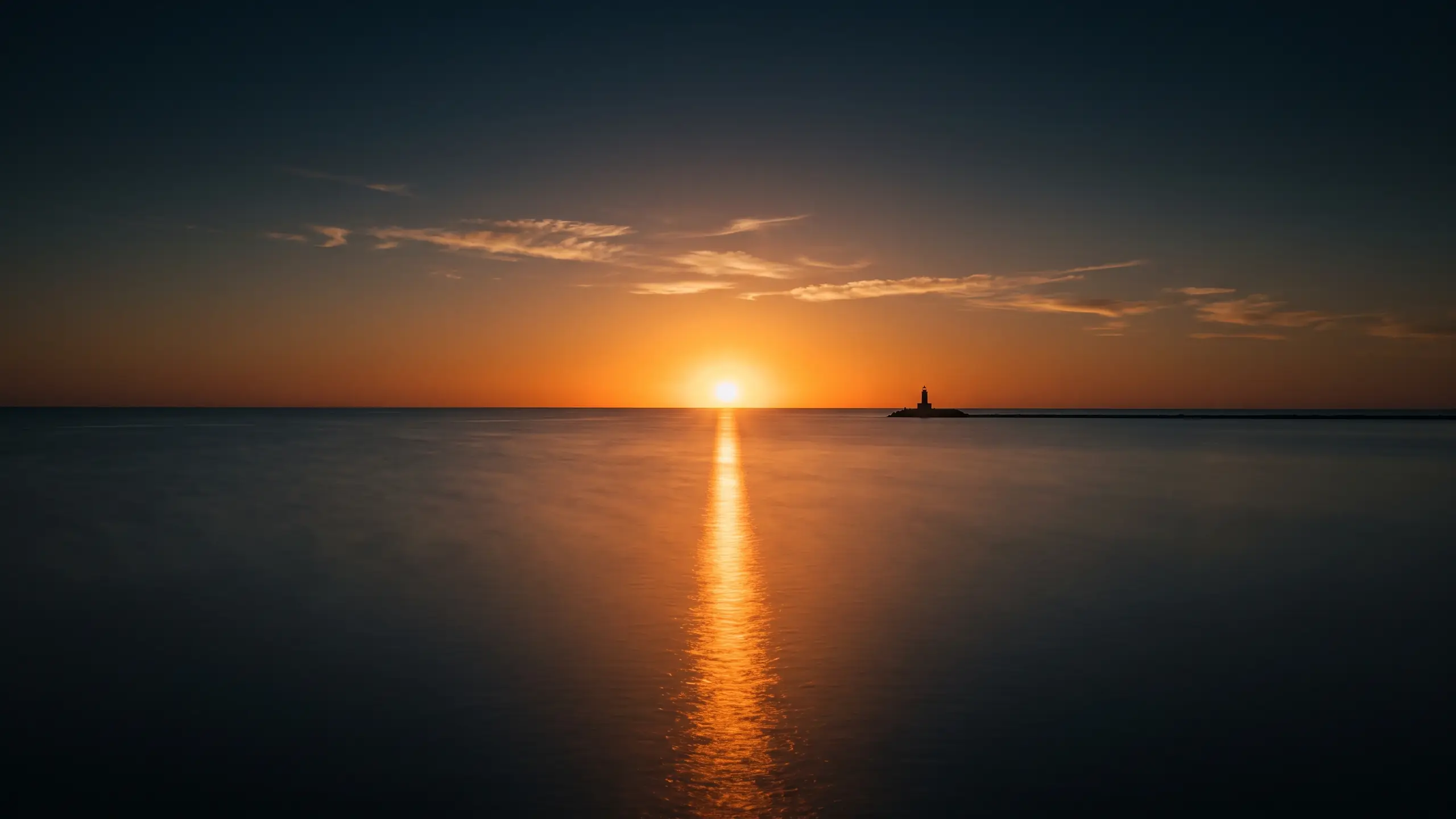 A calm lake at sunrise with an orange sun on the horizon, its reflection running toward the viewer, and a small lighthouse at the right edge
